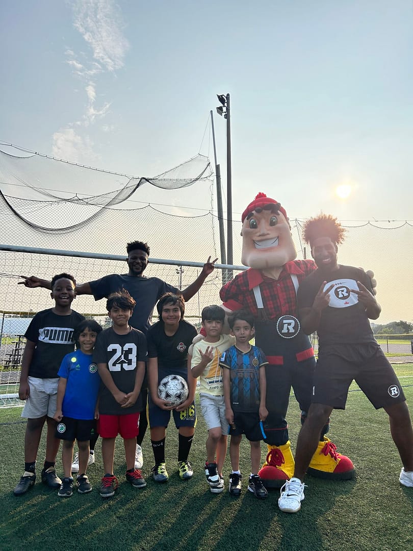 REDBLACKS mascot Big Joe, REDBLACKS athlete Alonzo Addae, joined alongside smiling with group of youth.